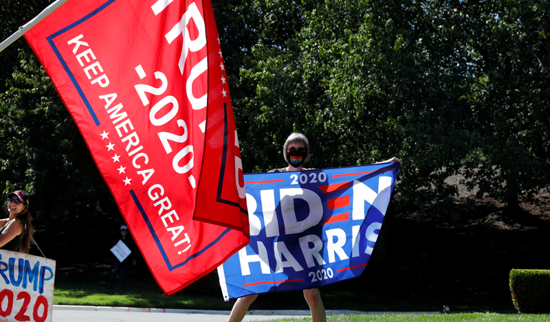 Photo of a woman holding up a "Biden Harris" sign. There is a "Trump 2020" sign in the foreground.