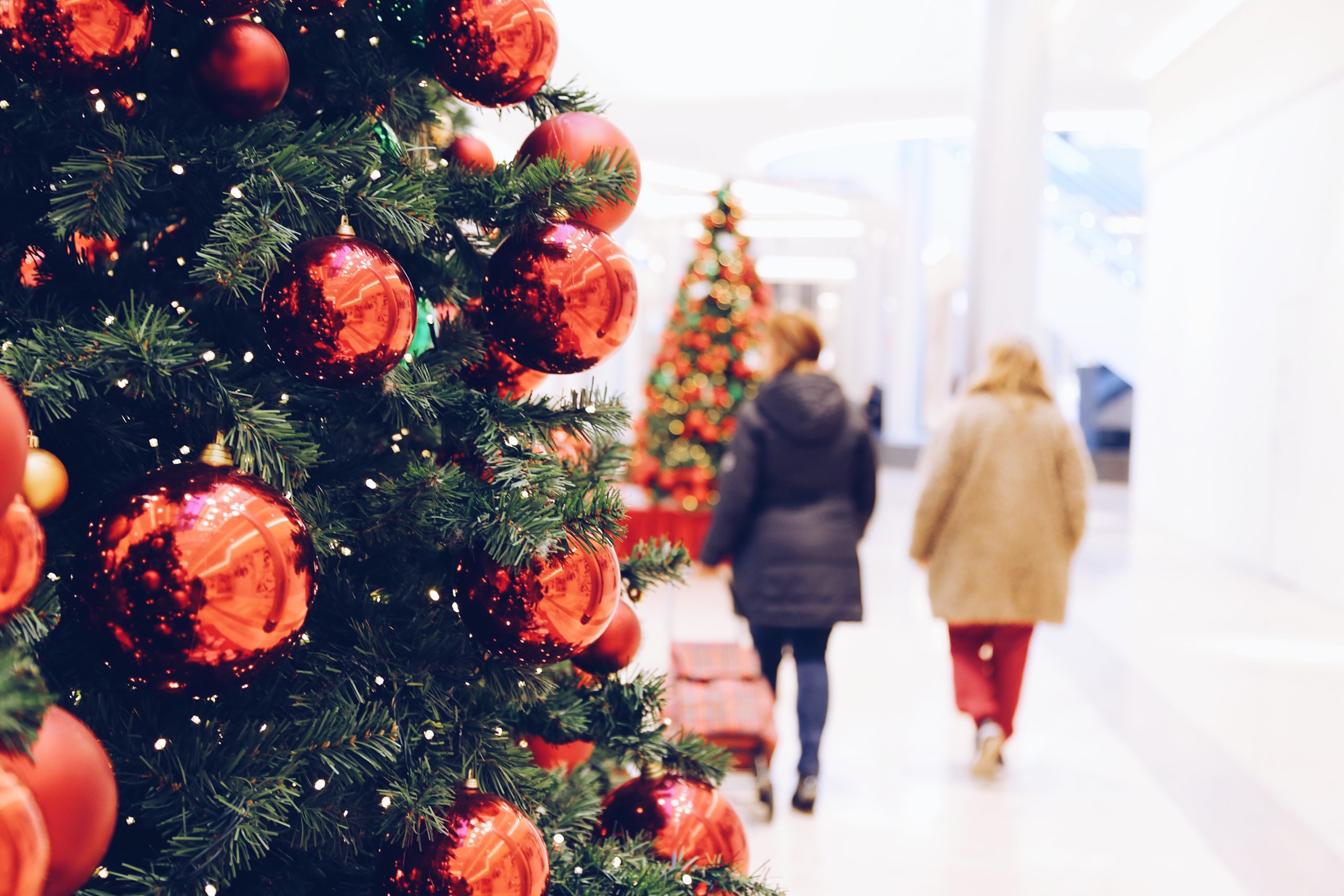 Photo of a Christmas tree and its ornaments. Two people are walking away from the camera's view. We see their backs.