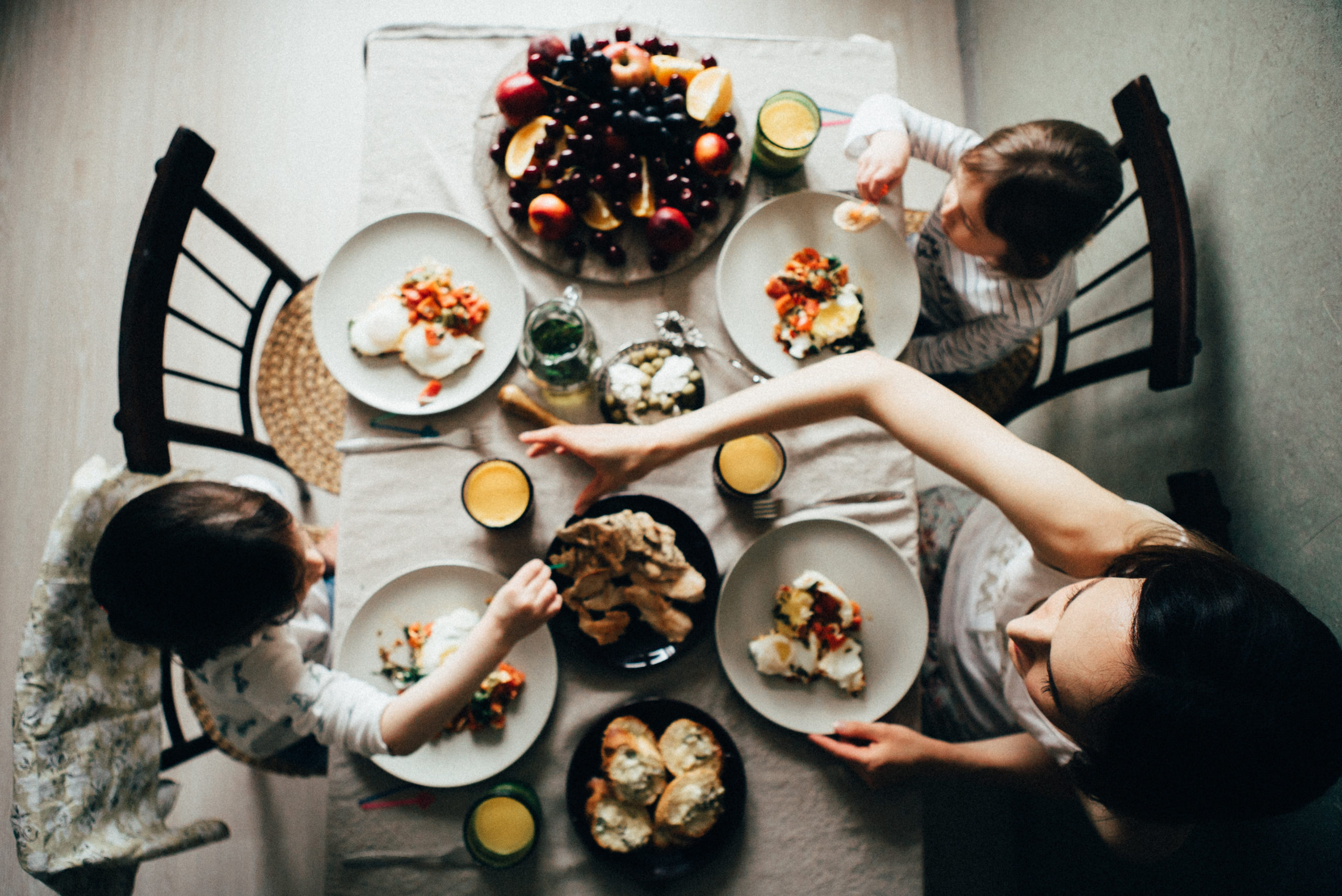 Aerial photo of two children eating breakfast with their mother. A fourth plate is set at the table but no one occupies its seat.