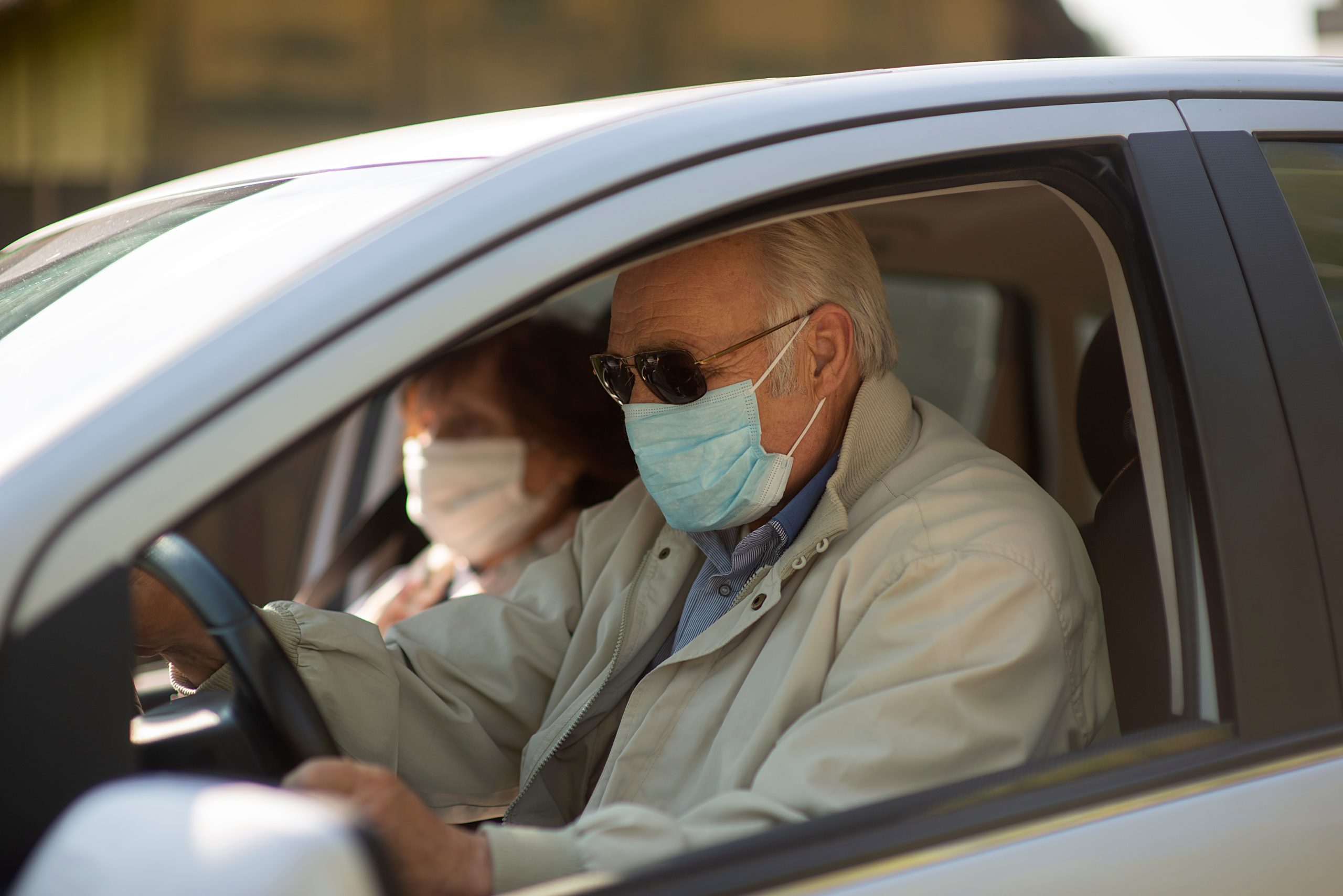 Photo of an elderly couple inside of a car. Both are wearing surgical masks.