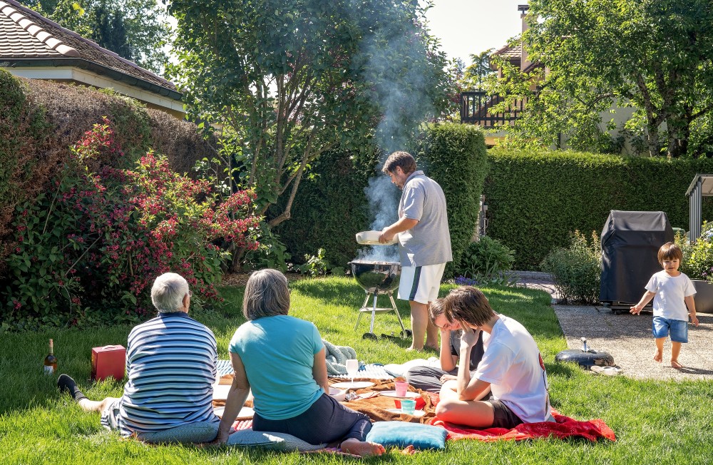 A group adults sit in a backyard while one of them barbeques.