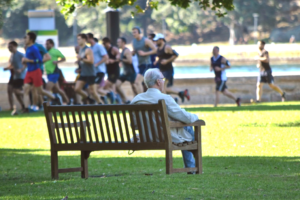 An older man sits on a bench in a park as a group of young runners go by.