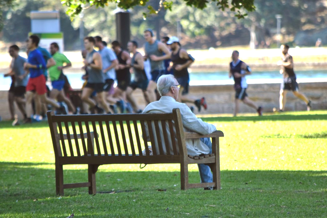 An older man sits on a bench in a park as a group of young runners go by.