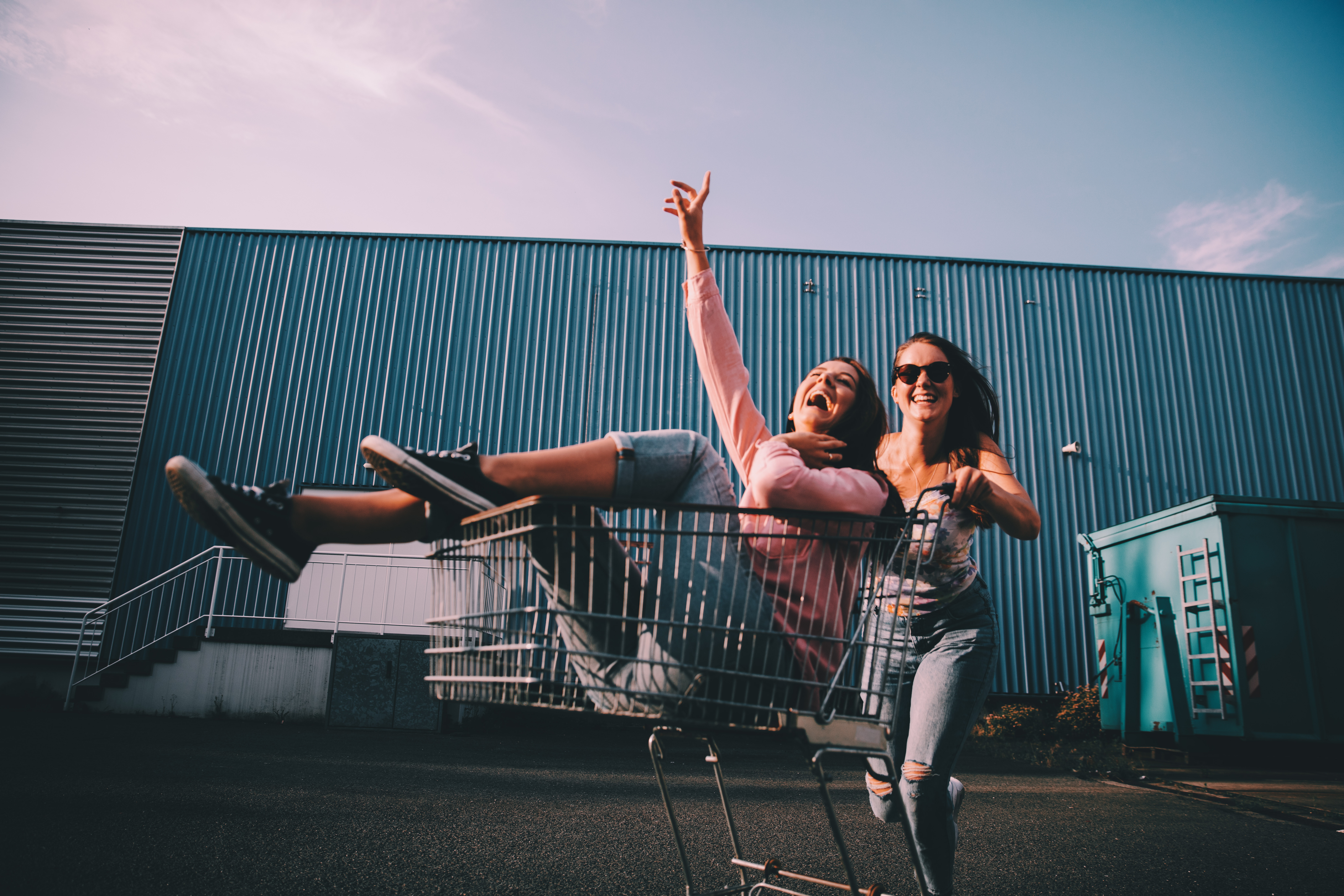 In a parking lot against a pale blue sky and dark blue metal wall, two young women laugh and smile while one sits inside of a shopping cart while the other pushes the cart.