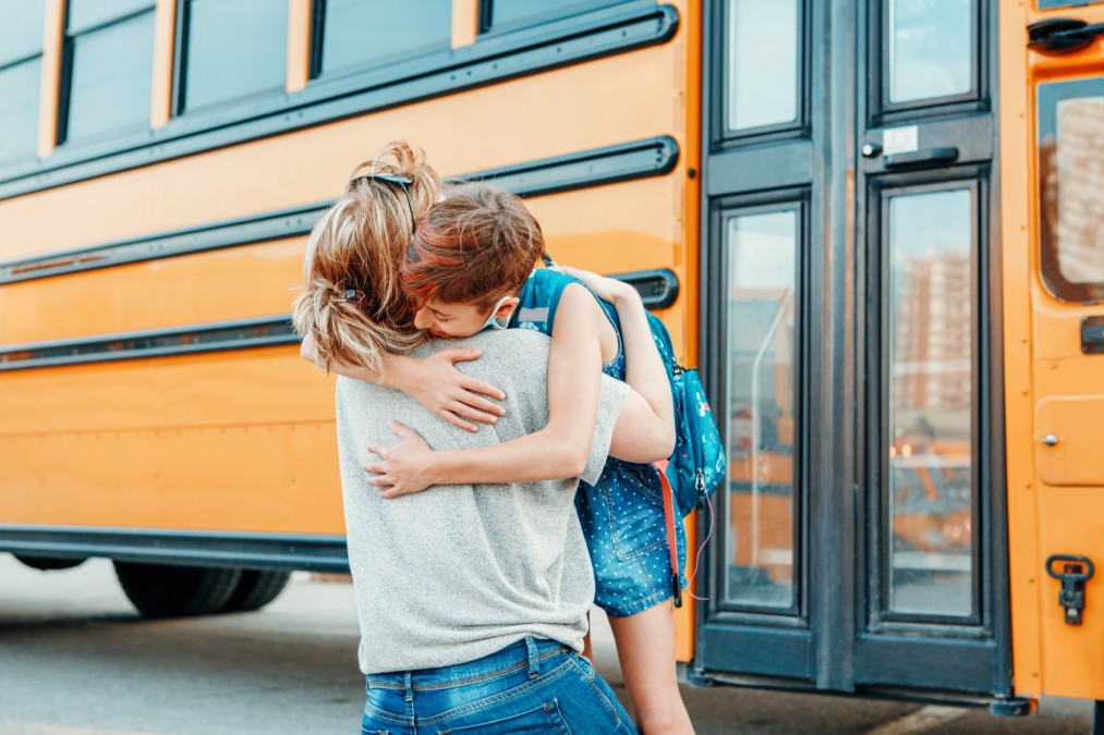 A Caucasian mother embraces her child on the school yard in front of a yellow school bus.
