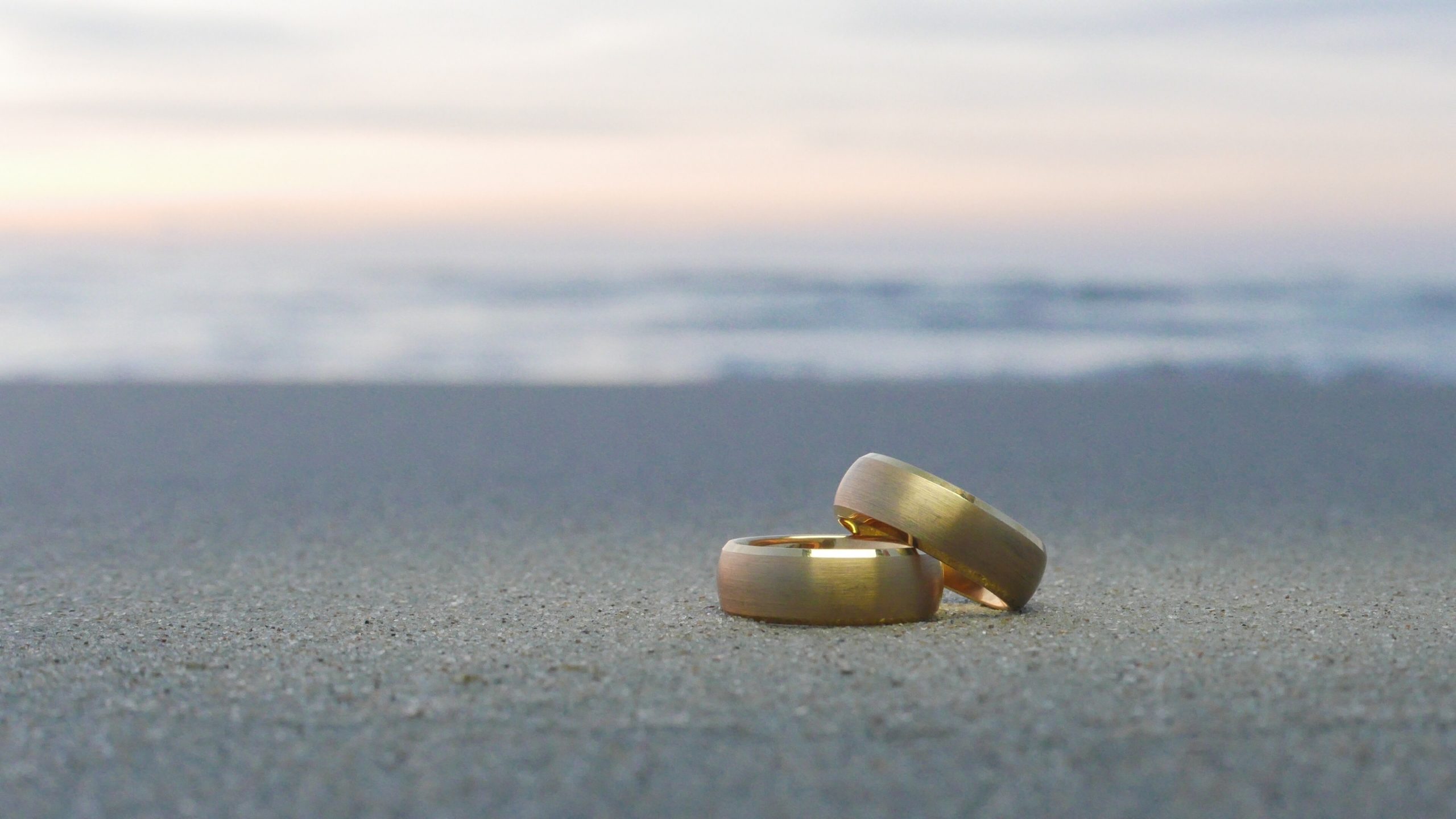 A pair of wedding rings rest on a sandy beach at sunset.