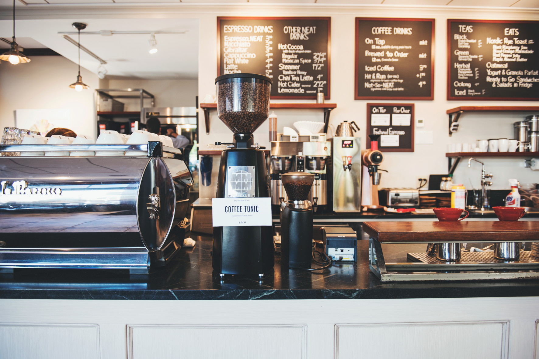 The bar of a coffee shop, including an espresso machine, espresso grinder, and blackboard listing a variety of drink offerings.