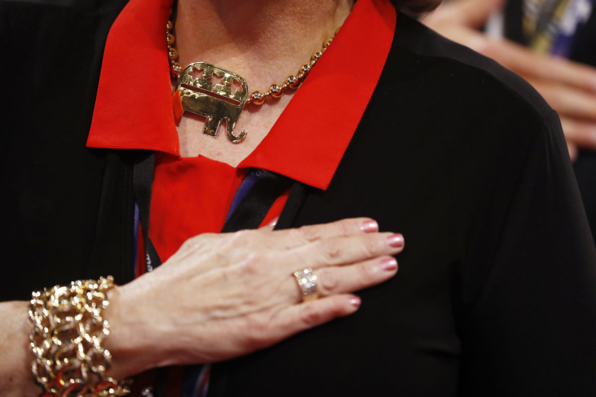 The upper torso of a white woman with her hand crossed over her heart. She is wearing a black jacket and red collared shirt. Around her neck is a large gold necklace of the Republican Party elephant.