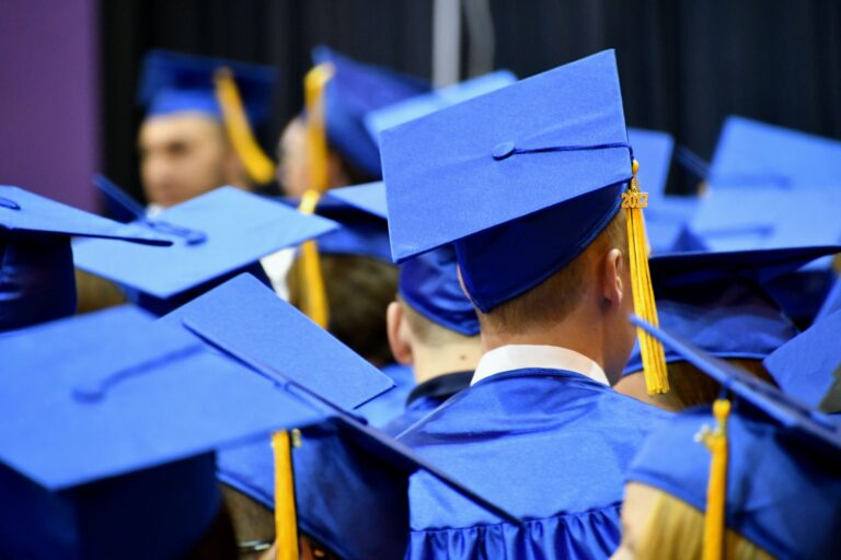 From behind, a close up of a single white male in a blue graduation cap and gown, surrounded by other graduates wearing the same caps and gowns.