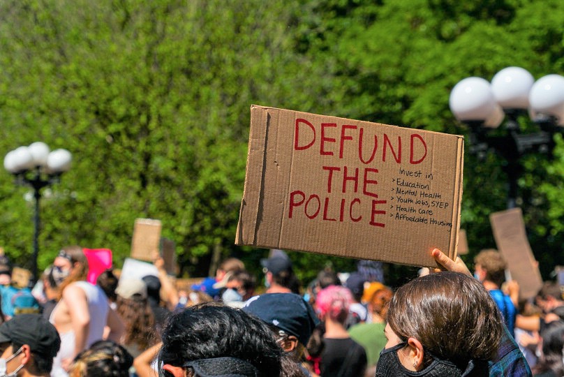 A white man holds a sign reading "DEFUND THE POLICE" in red lettering at a protest.