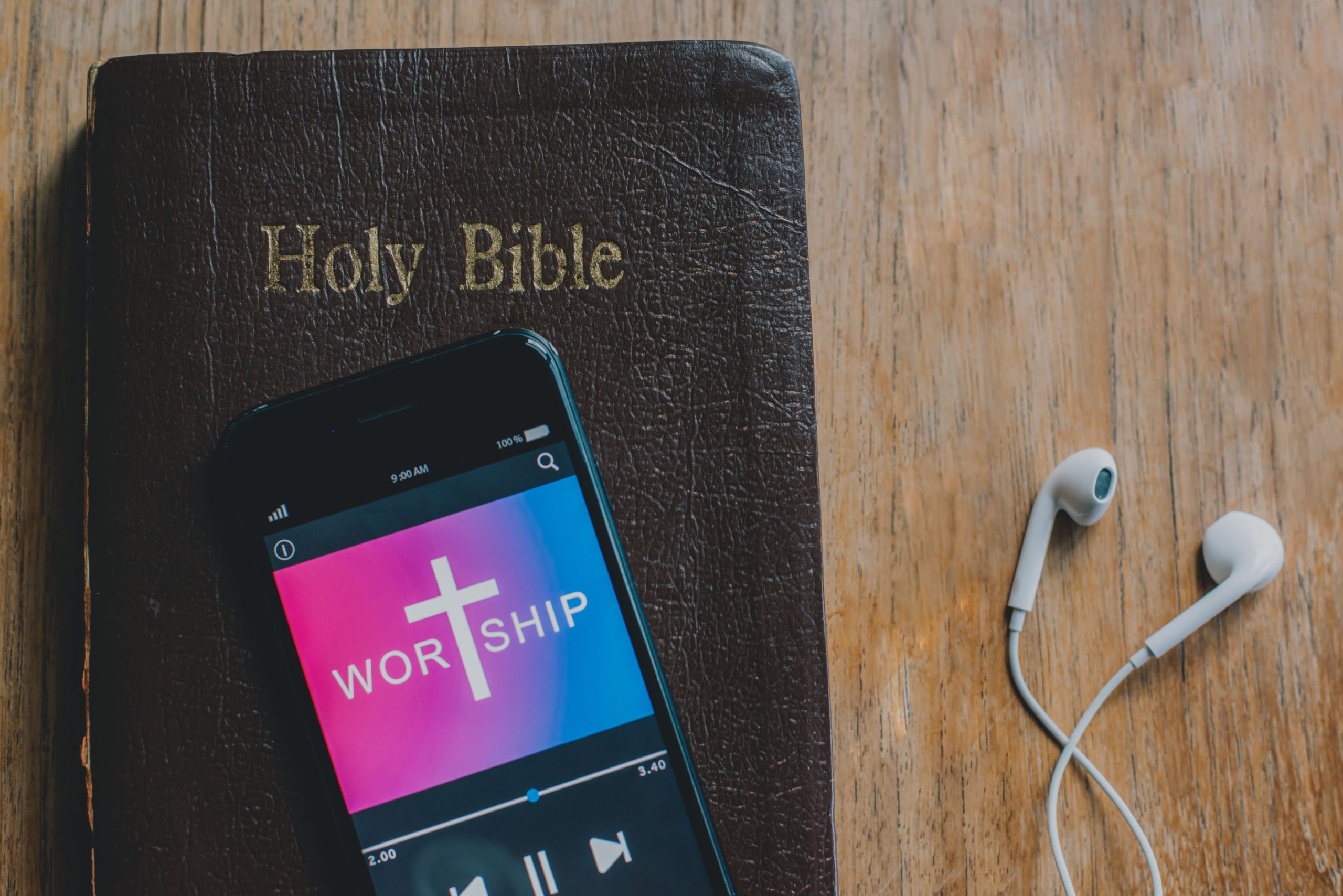 A photo of a phone playing worship music atop of the Bible. There are earbuds on the right side of the photo.