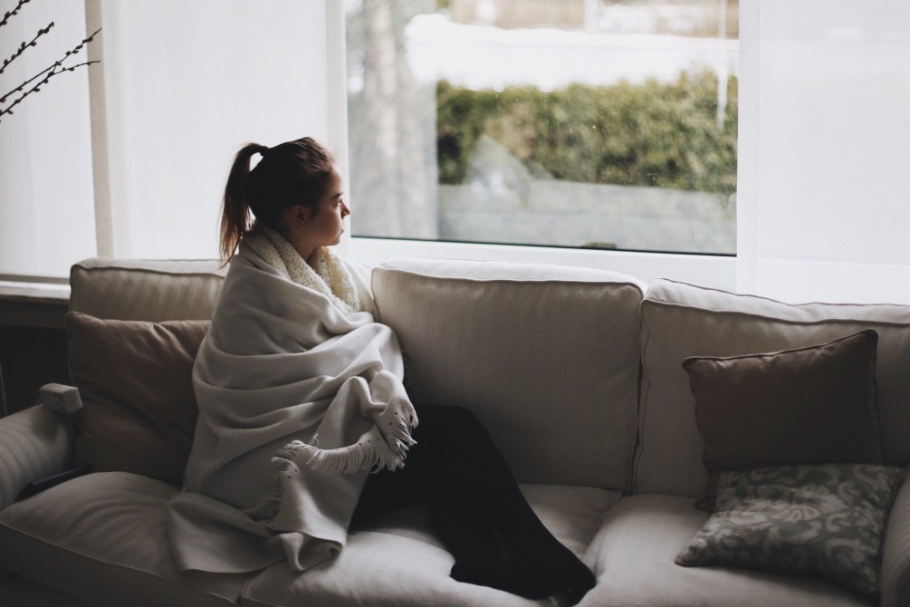 A young woman wrapped in a blanket stares pensively out the window.