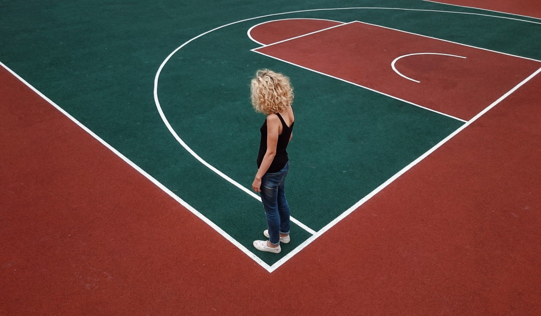 A young woman with curly blond hair stands alone on a red and green athletic court.