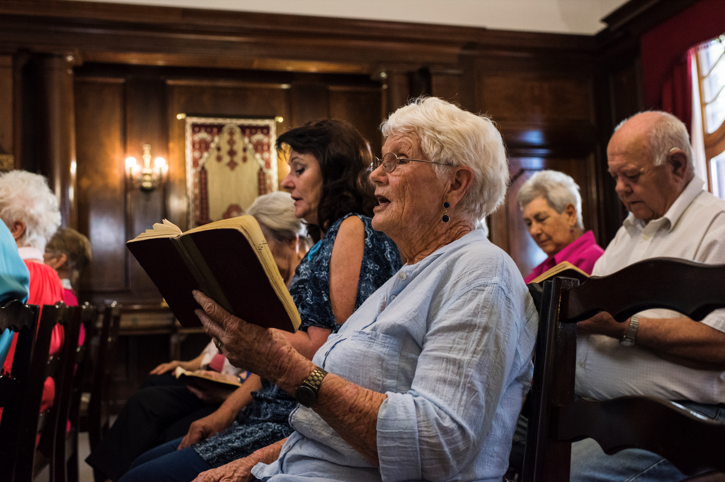 Grey haired woman singling hymn in pew sitting down in front of wood paneled wall and other congregants.