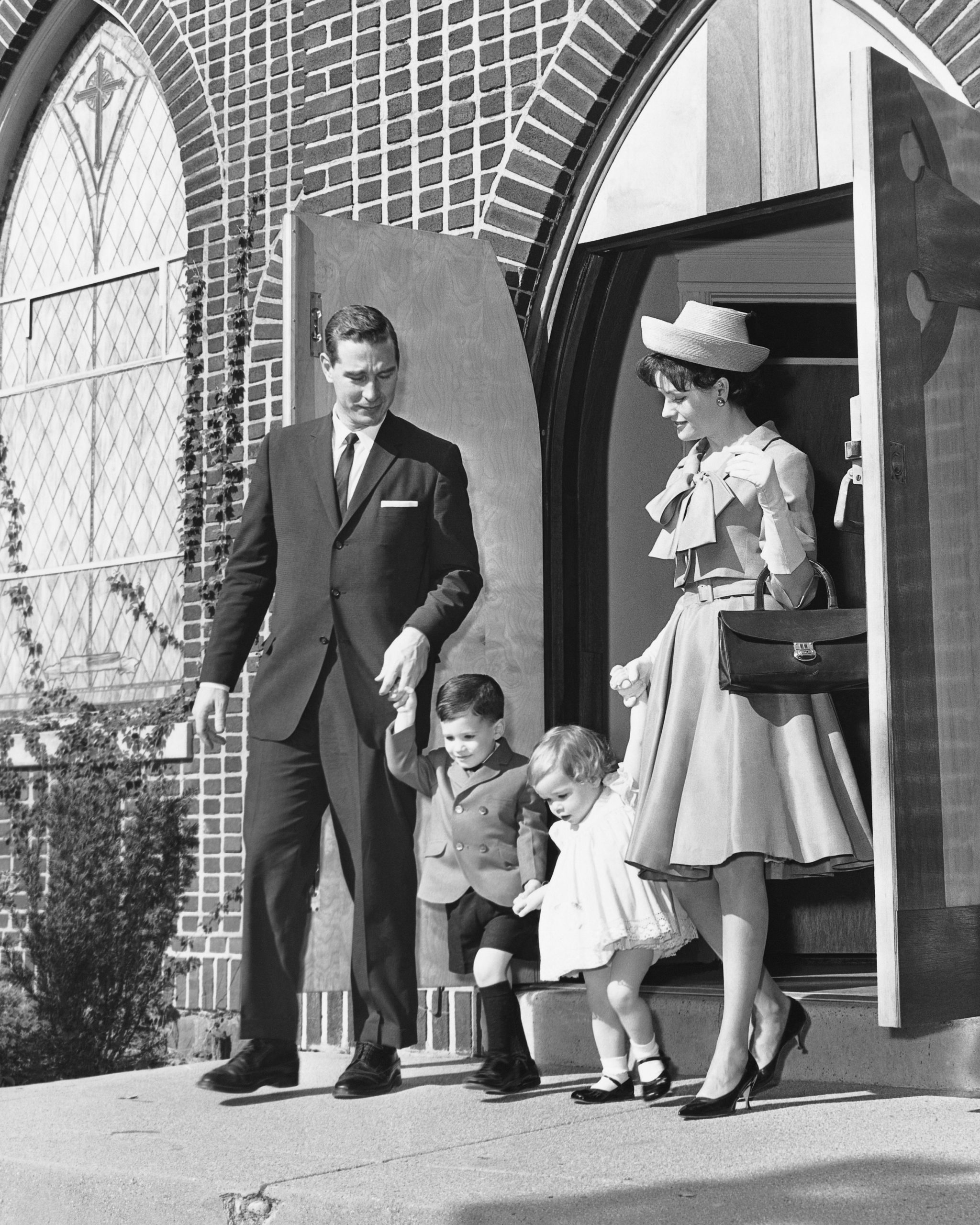 Black and white image of well dressed white married couple with young boy and girl exit church in 1950s.