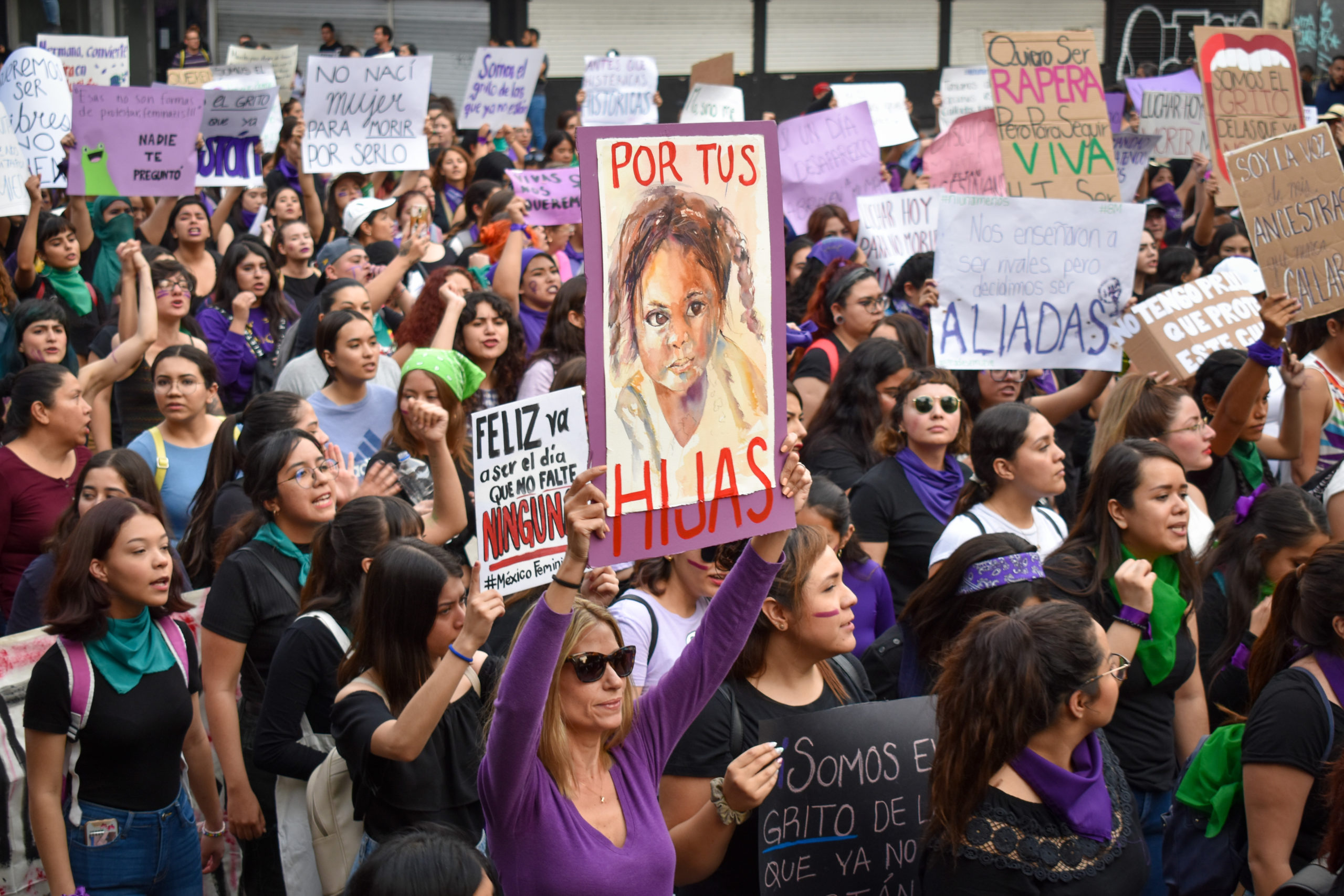 Large group of Hispanic women protesting in green bandanas, one wearing purple in focus in the forefront holds up a sign that says "por tus hijas" or "for your daughters" with a picture of a young girl on in.