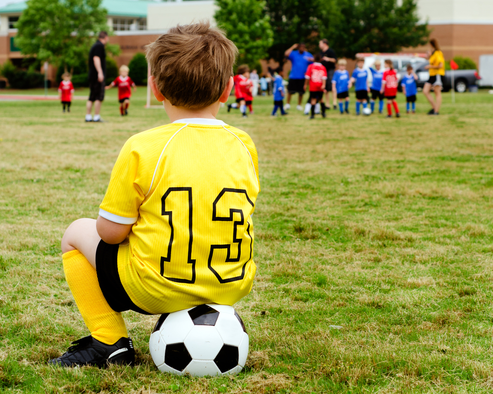 young boy sitting on a soccer ball alone on the wedge of a soccer pitch watching other kids play